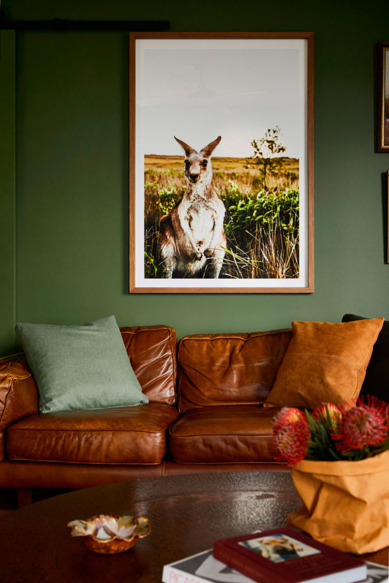 The living room is done with green walls, a brown leather sofa, a rich-toned wooden table and a bold artwork
