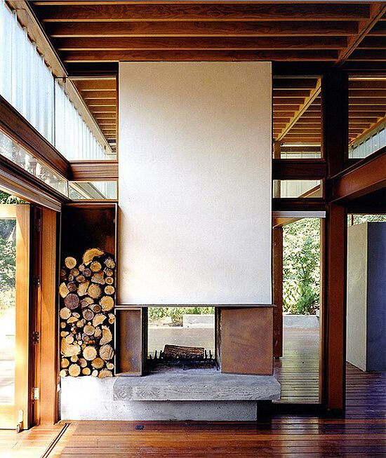 An ultra modern double sided fireplace with a white panel over it and metal parts plus firewood stored next to it