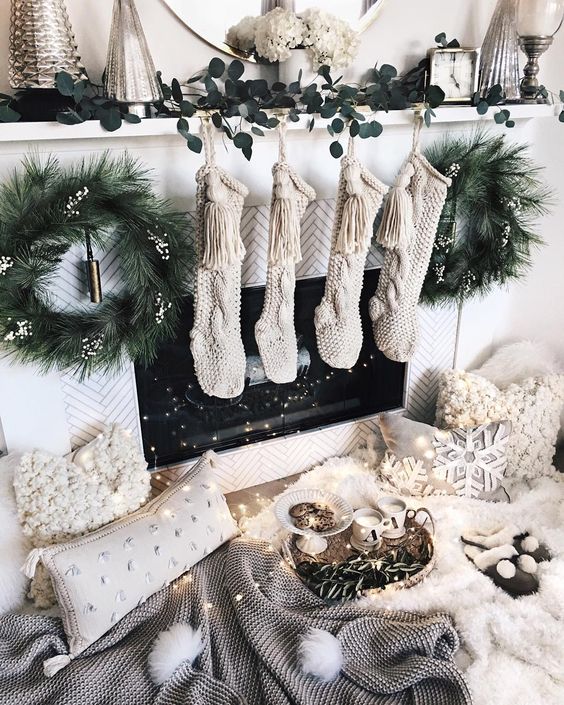 a magical fireplace with knit stockings, evergreen wreaths, a greenery runner and fluffy pillows and lights