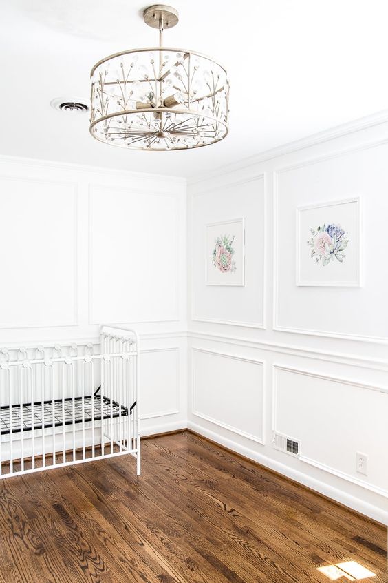 A gorgeous nursery with paneling, warm colored hardwood floors and a beautiful chandelier