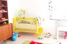 a bright and fun nursery with a printed rug, a yellow baby crib, colorful pompom garlands and bright toys