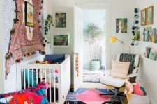a bright boho nursery with a pompom rug on the wall, colorful rugs on the floor, pillows and bright bedding