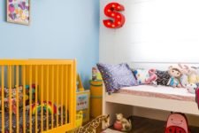 a brigth nrusery with a yellow crib, a printed rug, a marquee letter, bright toys and artworks for fun