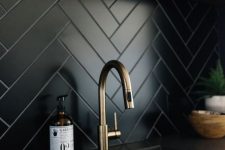a chic dark kitchen with a matte black herringbone backsplash, black coutnertops, a floating shelf and brass touches