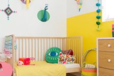 a colorful nursery with a color block wall, a printed rug, colorful mobiles and fun art on the wall plus brigth bedding