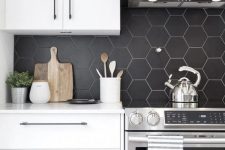 a contemporary kitchen with white cabinetry, a black matte hexagon tile backsplash and a black hood looks contrasting