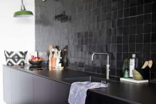 a cool black kitchen with sleek lower cabinets, tiles all over the wall, green lamps and stone countertops