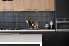 a minimalist black kitchen with stained upper cabinets, a black skinny tile backsplash, a white kitchen island and a black pendant lamp
