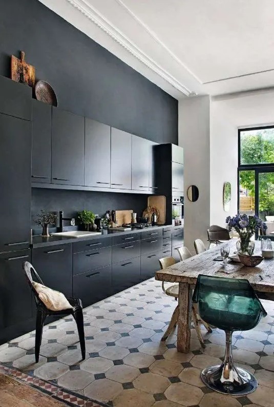 a stylish black kitchen with a matte black wall, black countertops and a backsplash, a rough wooden table and mismatching chairs