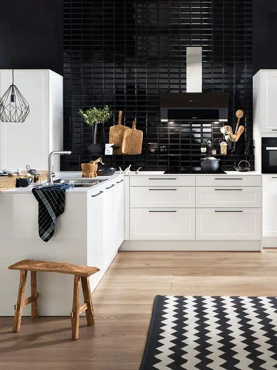 A stylish modern white kitchen of an L shape, with a glossy black tile backsplash and a wooden stool, some greenery and a chevron rug