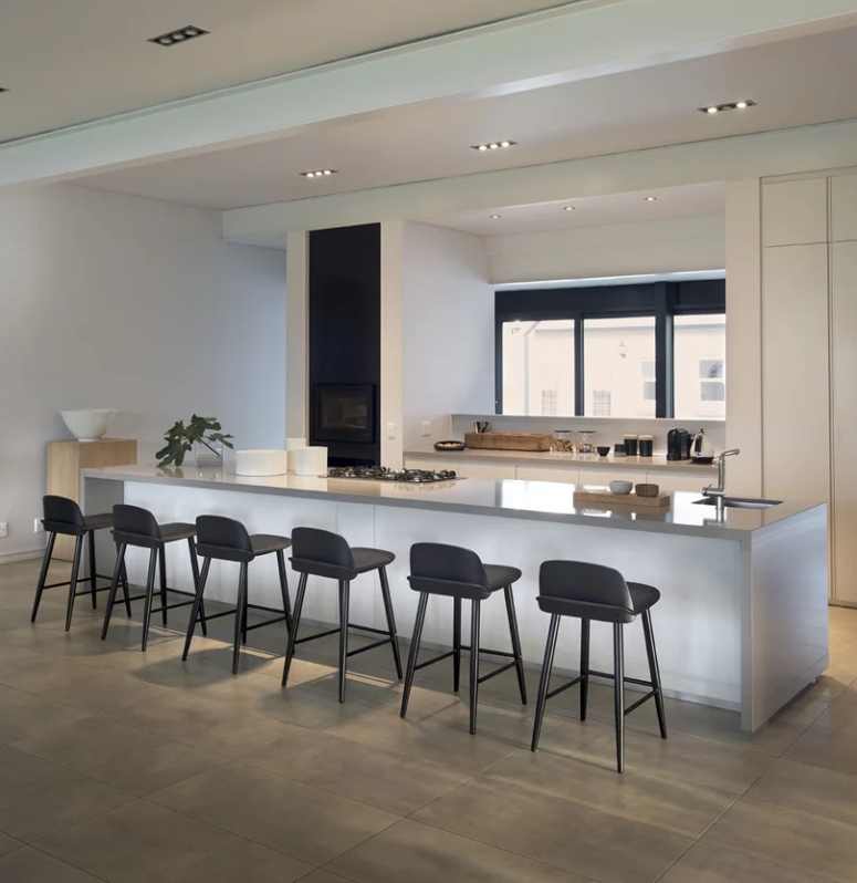 The kitchen is all-white and sleek, with a giant kitchen island, a window to bring light in and black stools