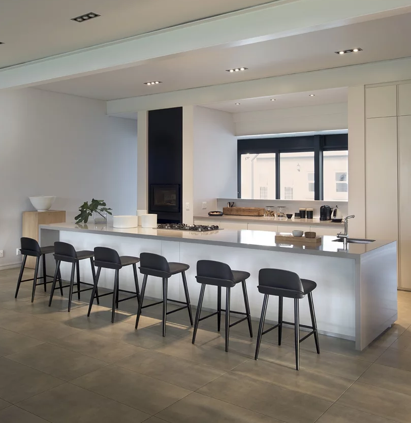 The kitchen is all white and sleek, with a giant kitchen island, a window to bring light in and black stools