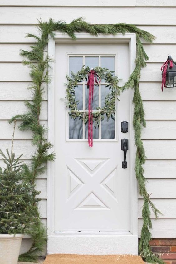 a simple evergreen garland covering the doorway, mini Christmas trees in pots and a greenery wreath with a red ribbon bow