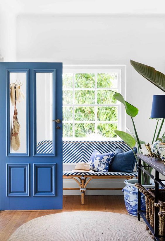 A stylish entryway dotted with classic blue   a door, a lamp, pillows, a striped upholstered bench and patterned pot