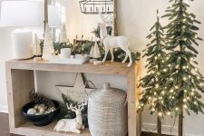 a farmhouse Christmas console with deer figurines, a basket with a lid, evergreens and lights and a duo of Christmas trees