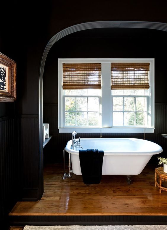 a moody bathroom with black walls, Roman shades, a wooden platform and a white bathtub