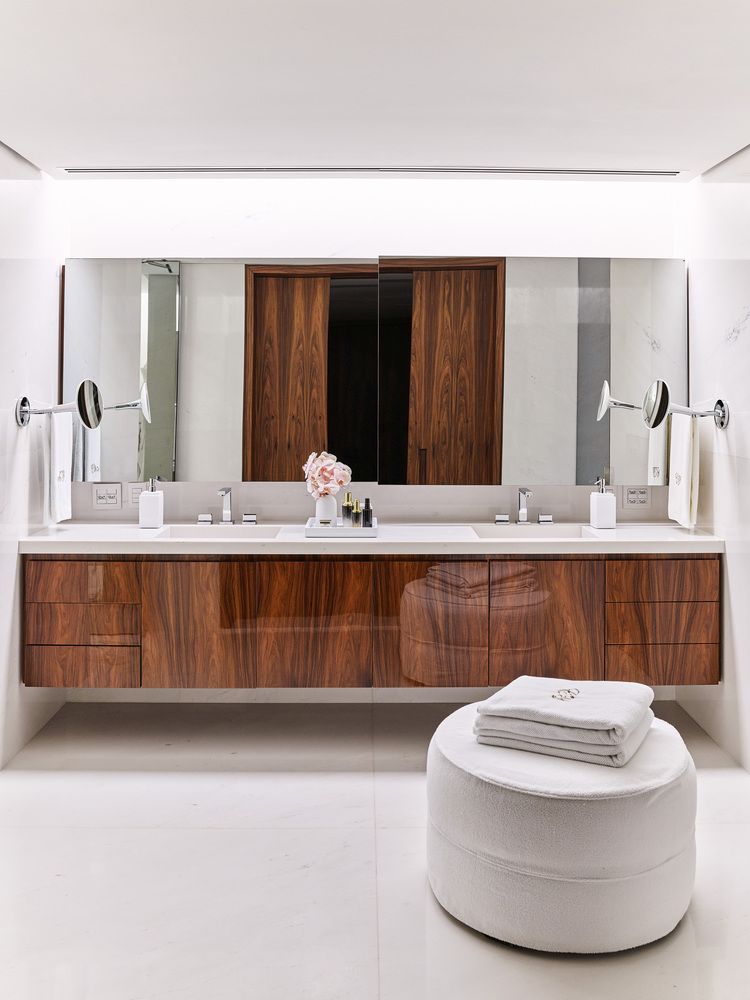 The bathroom is done with white stone, a wooden vanity and a large comfy ottoman