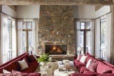 a chalet living room with a wooden ceiling with beams, a stone fireplace, red sofas and whitewashed coffee tables