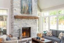 a clean double-height neutral living room with a grey stone fireplace with a wooden mantel and a bright blue artwork