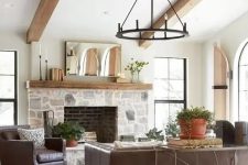 a modern farmhouse living room with a stone clad fireplace, wooden beams, leather furniture, a metal chandelier and potted plants
