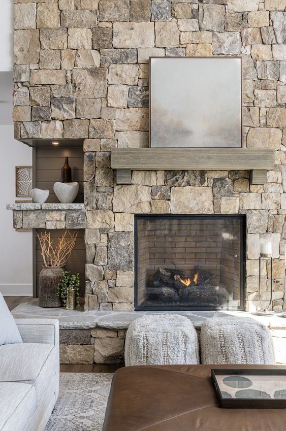a modern living room with a stone fireplace and a mantel, some niche shelves and seating furniture and a leather ottoman