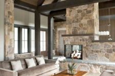 a neutral contemproary cabin living room with a stone double-sided fireplace, neutral furniture, wooden beams