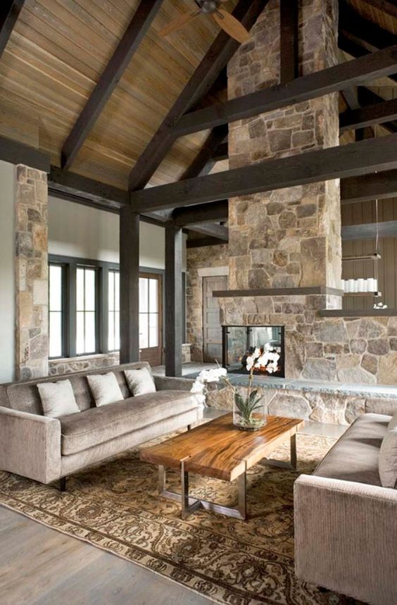 a neutral contemproary cabin living room with a stone double-sided fireplace, neutral furniture, wooden beams