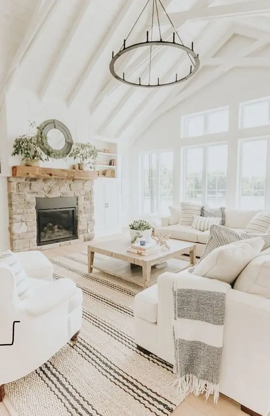 A neutral farmhouse living room with wooden beams, a round chandelier, white furniture, a built in fireplace in stone