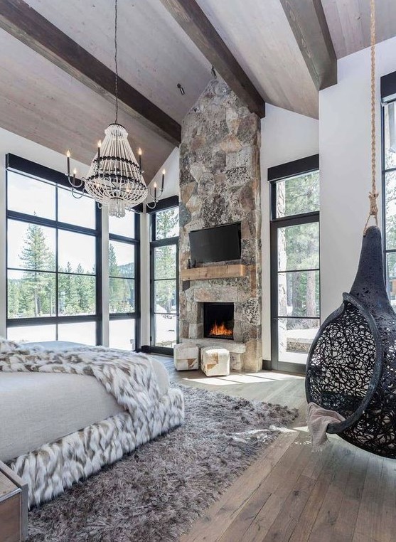 An ultra modern chalet bedroom with a stone fireplace, a suspended chair, a catchy chandelier and lots of faux fur