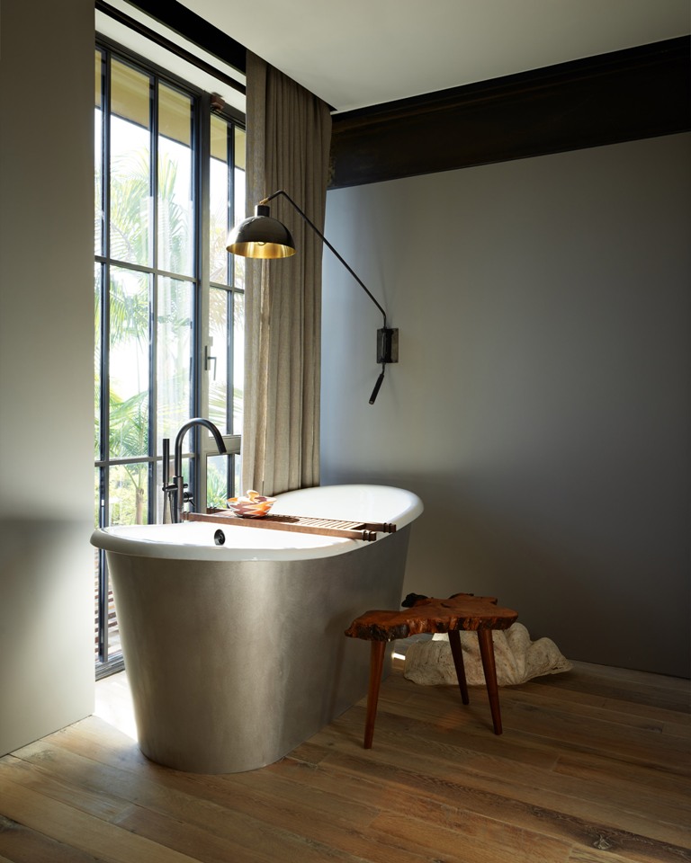 The bathroom is clean and features a full height window with a curtain, a silver oval tub and a catchy wall lamp