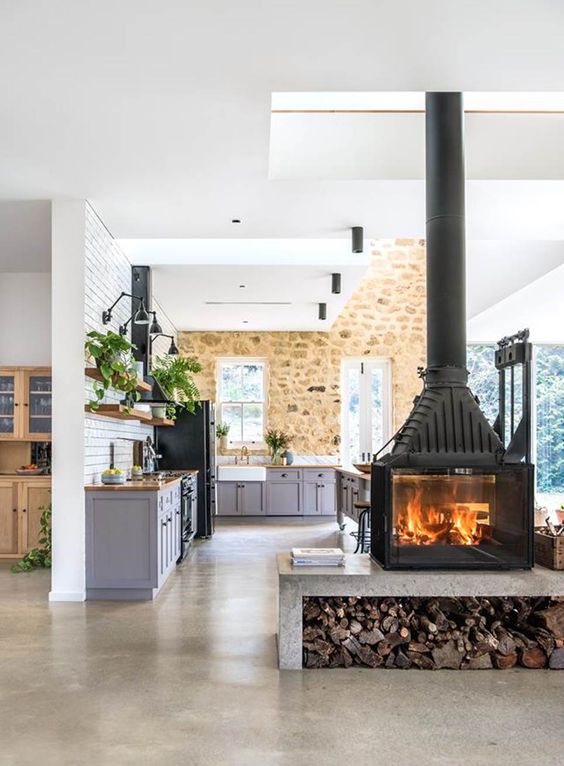 An oversized wood burning stove on a concrete platform with firewood divides the spaces in a cozy way