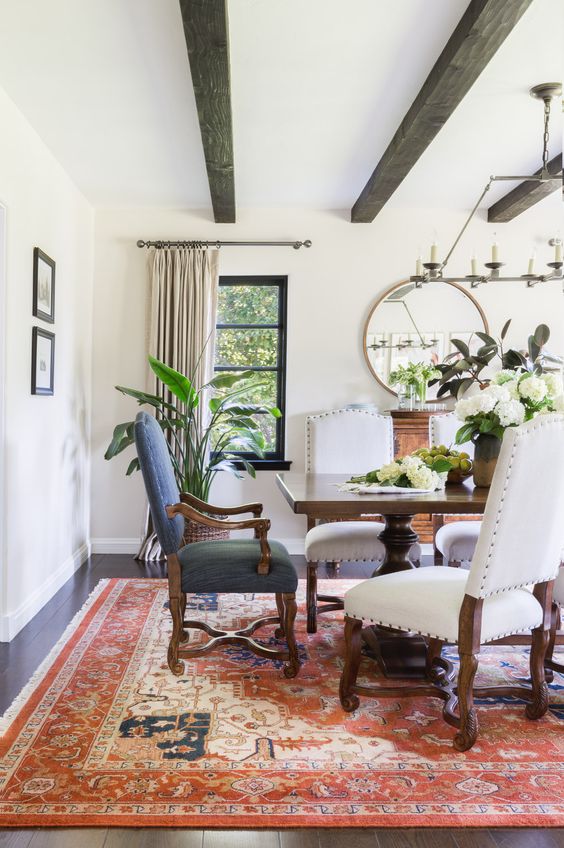 a Spanish dining room with heavy dark wooden furniture, a printed rug, a round mirror and lots of greenery and blooms