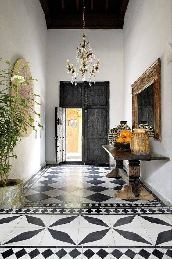 a Spanish entryway with black and white printed tiles, a reclaimed dakr wood door, a heavy console and potted greenery