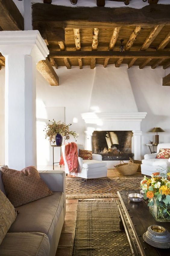 a Spanish living room with a wooden ceiling with beams, white walls and a fireplace, neutral furniture and dried blooms