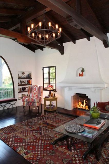 a Spanish living room with white plaster walls, a dark ceiling with beams, a large fireplace and pritned textiles and upholstery