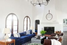 a Spanish living room with white walls and a double-height ceiling, dark wooden beams, a large fireplace and jewel tone furniture