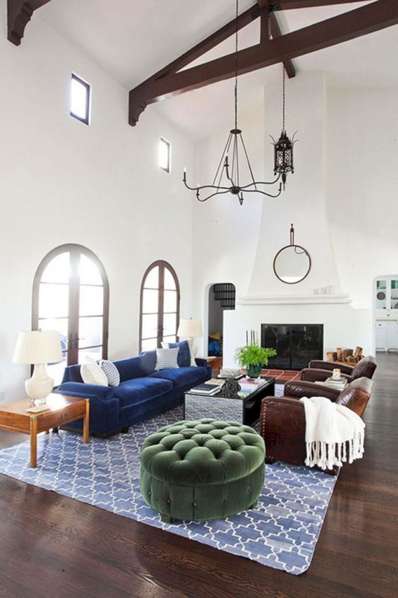A Spanish living room with white walls and a double height ceiling, dark wooden beams, a large fireplace and jewel tone furniture