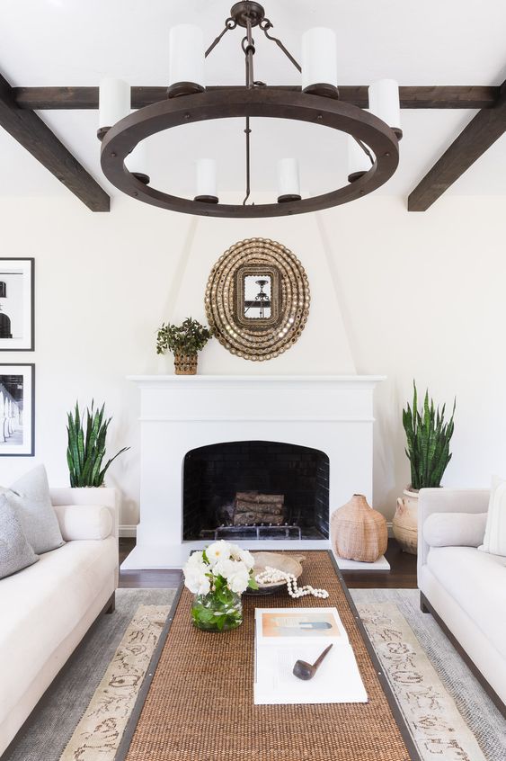 a Spanish living room with white walls, dark wooden beams, a matching chandelier, a fireplace and natural touches