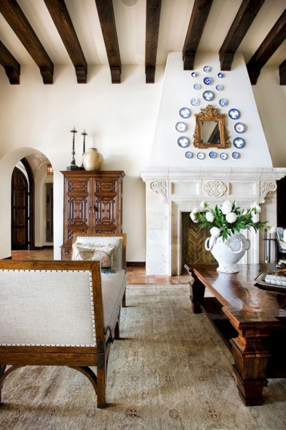 a Spanish living room with white walls, tiled floors, dark wooden beams, elegant and heavy furniture and a large fireplace