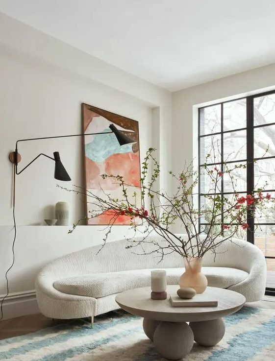 a beautiful living room with a curved creamy sofa, a round coffee table, a bold artwork, black wall lamps and a glazed wall