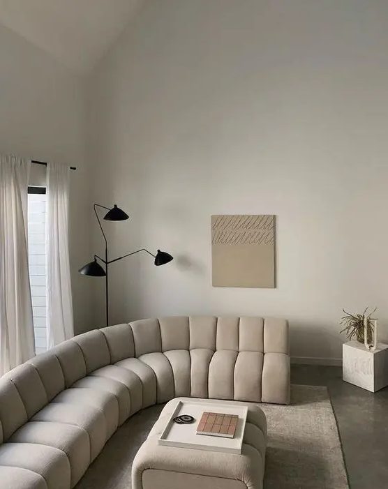 A beautiful warm colored neutral living room with a creamy curved sofa and a matching pouf, a black floor lamp and a cube
