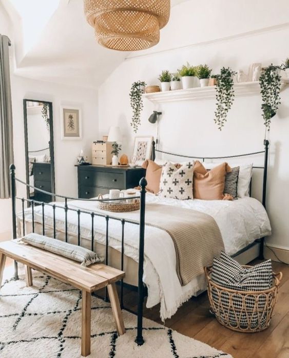 a boho bedroom with a forged bed, potted greenery, a wicker lamp, wooden furniture and dark touches