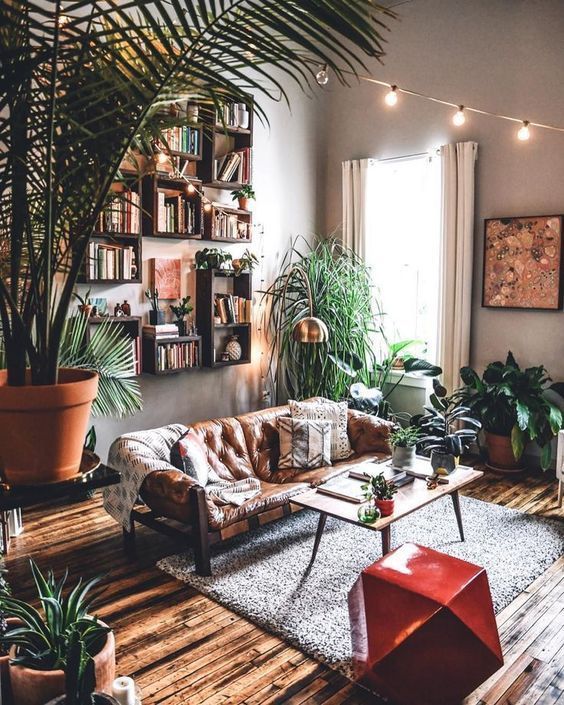 a boho living room with an arrangement of bookshelves, a leather sofa, lots of potted greenery and a geometric stool