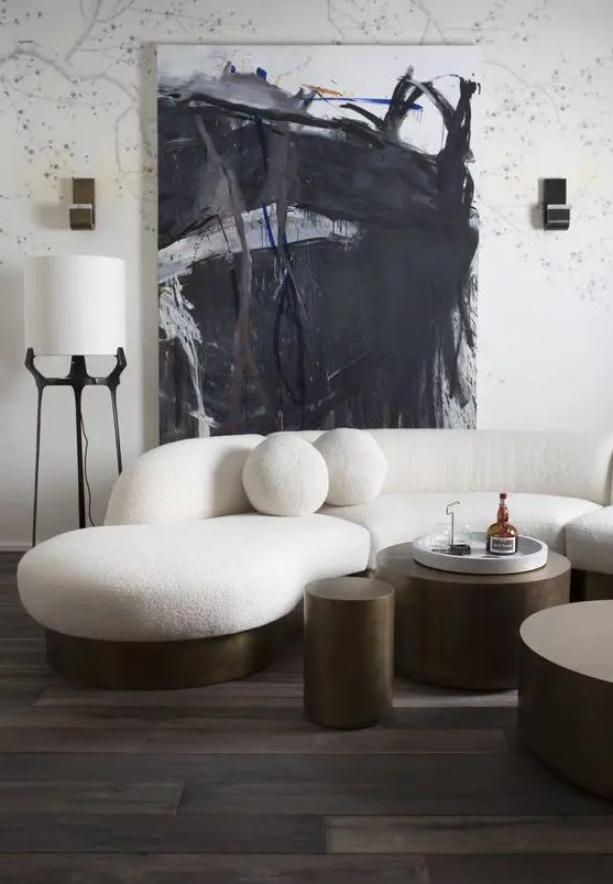 a bold and catchy living room with a white curved sofa with round pillows, metal tables, an abstract artwork and some lamps
