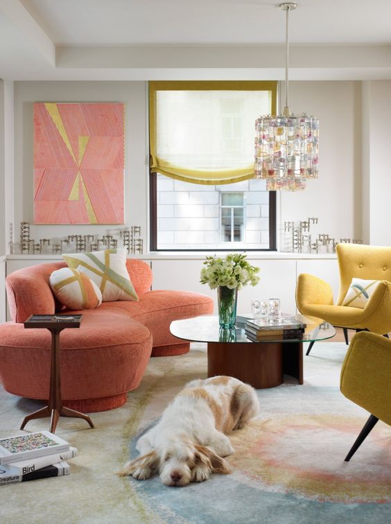 a bright living room with a coral curved sofa, yellow chairs, a pastel rug, a coffee table and a bright chandelier