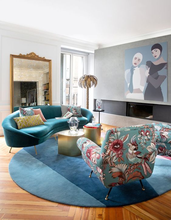 A colorful and eclectic living room with a built in fireplace, a large artwork, a blue curved sofa and a rug, floral print chair and gold coffee tables