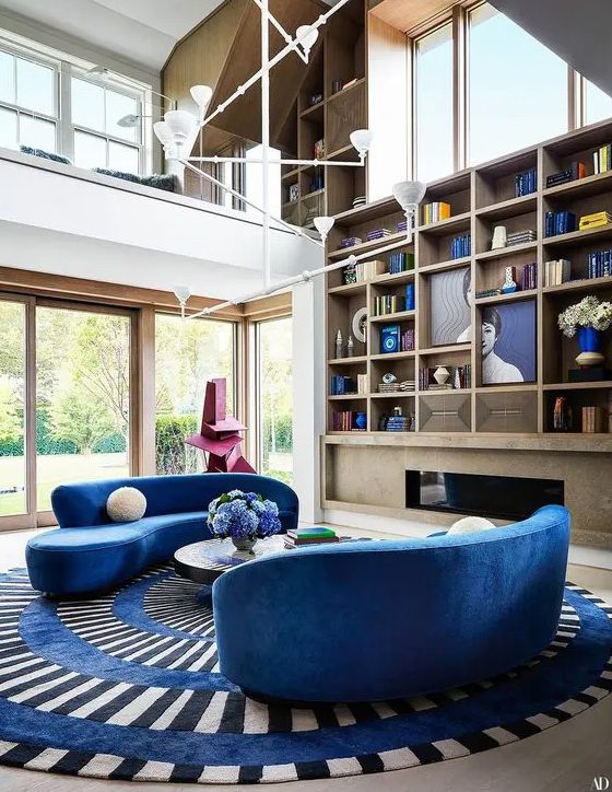 A colorful living room with a built in fireplace, a large built in bookcase, bright blue curved sofas, a striped rug and a coffee table