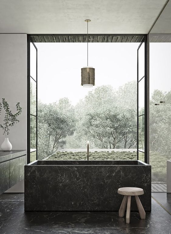 a gorgeous moody bathroom with a black square stone bathtub and matchign floors and a vanity plus a garden view