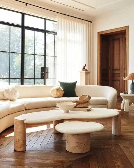 A light filled living room with a parquet floor, a curved neutral sofa, a coffee table and a stool and some lamps