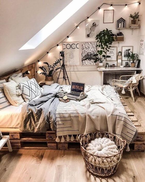 a neutral attic boho space with string lights, greenery in pots, a pallet bed, wicker and rattan furniture